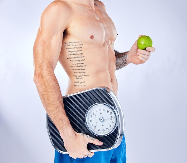 Báscula corporal y manzana con un hombre en el estudio sobre un fondo gris para una dieta de pérdida de peso o una alimentación saludable Tatuaje de fitness y salud con un hombre sosteniendo fruta mientras posa para un estilo de vida nutricional