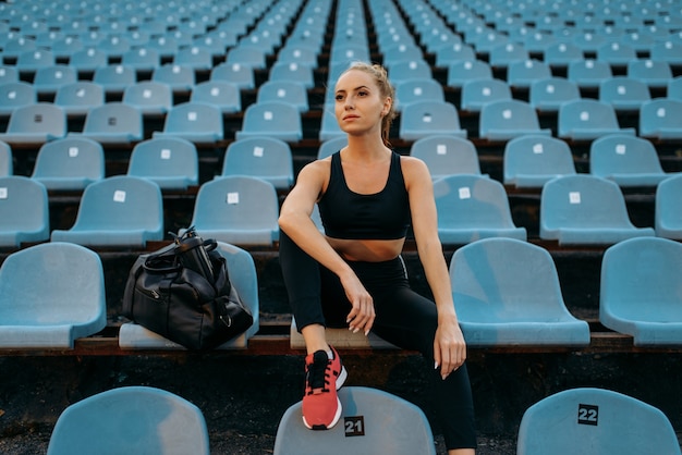 Basculador femenino en ropa deportiva sentado en la tribuna, entrenando en el estadio. Mujer haciendo ejercicio de estiramiento antes de correr en la arena al aire libre