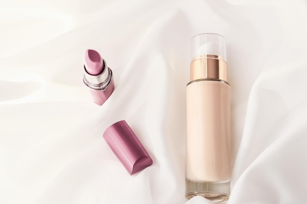 Base de base líquida de maquillaje de botella de crema tonal beige y lápiz labial rosa sobre productos cosméticos de fondo de seda como diseño de vacaciones de marca de belleza de lujo