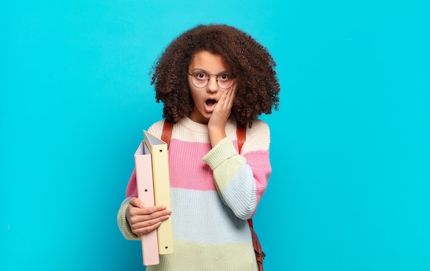 Bastante adolescente afro se siente conmocionado y asustado, mirando aterrorizado con la boca abierta y las manos en las mejillas. concepto de estudiante