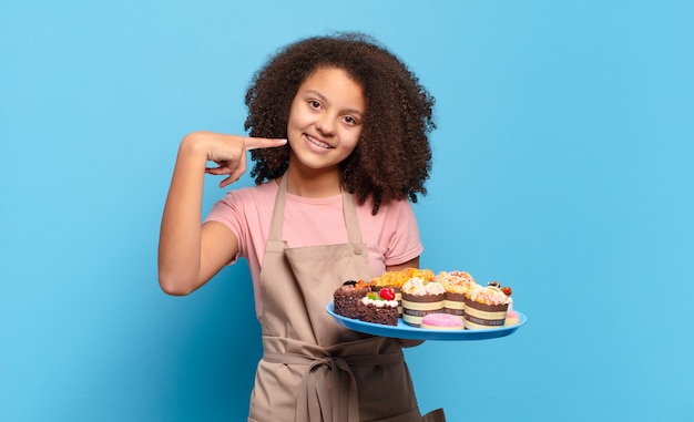 Bastante adolescente afro sonriendo con confianza apuntando a su propia sonrisa amplia, actitud positiva, relajada y satisfecha. concepto de panadero humorístico