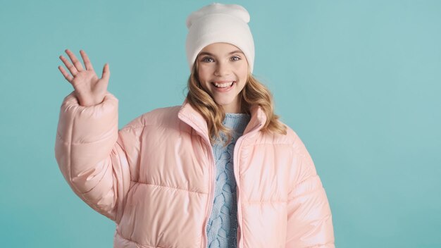 Bastante adolescente rubia vestida con chaqueta rosa y sombrero saludando a un amigo sobre fondo azul. Chica encantadora saludando hola