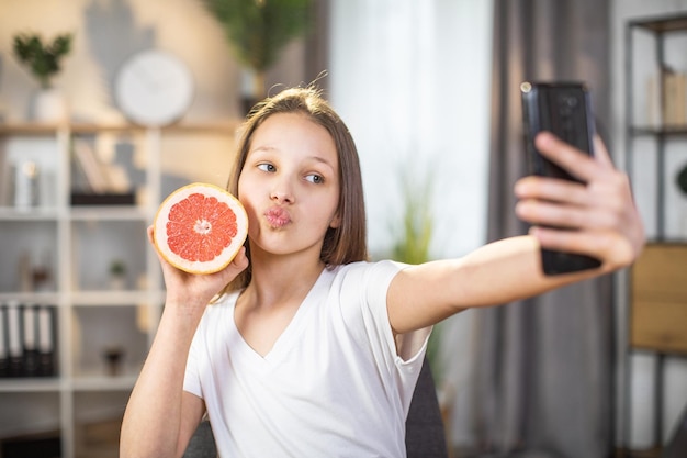 Bastante adolescente sosteniendo pomelo cerca de la cara y tomando selfie