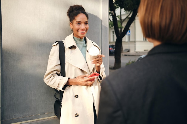 Bastante alegre chica afroamericana en elegante abrigo de trinchera sosteniendo café y teléfono celular mientras habla felizmente con un amigo al aire libre