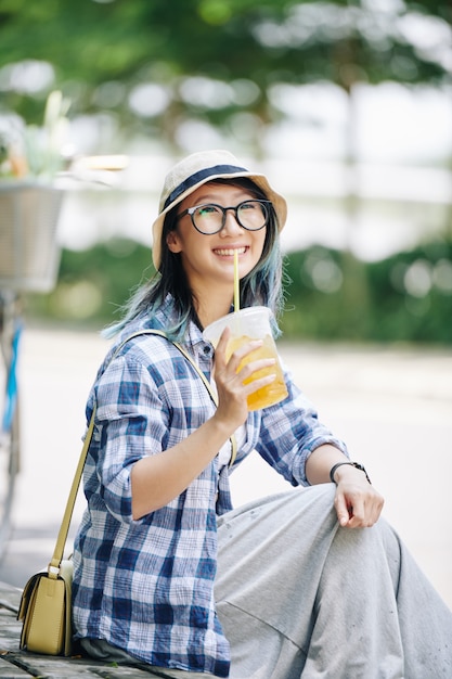 Bastante alegre joven China en vasos descansando al aire libre y bebiendo cócteles refrescantes