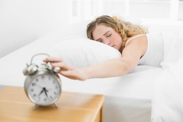 Bastante cansada rubia acostada en la cama apagando el despertador