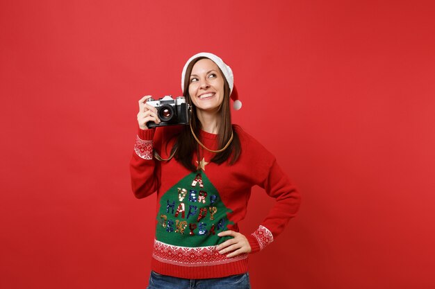 Bastante feliz joven Santa con sombrero de Navidad mirando hacia arriba y sosteniendo la cámara de fotos vintage retro aislada sobre fondo rojo. Feliz año nuevo 2019 celebración concepto de fiesta navideña. Simulacros de espacio de copia.