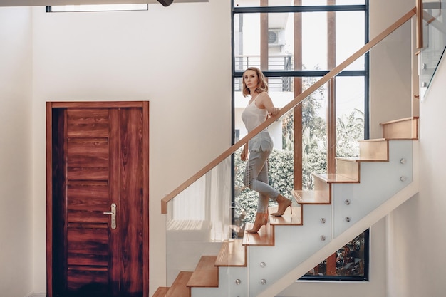 Bastante flaca y atractiva modelo femenina bajando una escalera de madera contra el fondo de una gran ventana. Concepto de vida rica