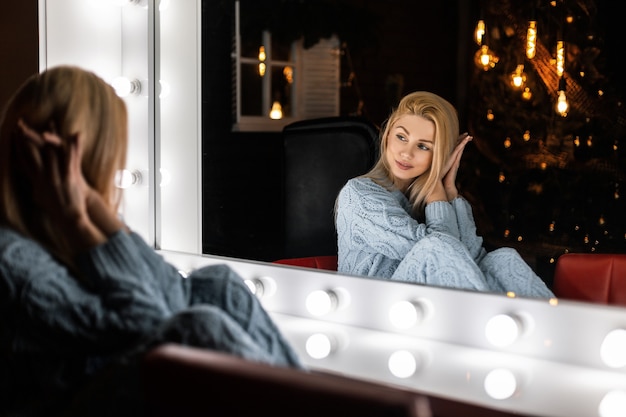 Bastante glamour joven con cabello rubio se sienta en una habitación con un árbol de año nuevo con luces brillantes y se ve en un espejo vintage con lámparas blancas