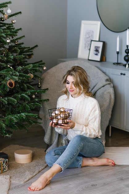 Bastante joven en un acogedor suéter y jeans decora un árbol de Navidad con globos