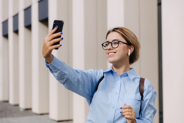 Bastante joven en anteojos de pie en la calle y con videollamada en el teléfono inteligente. Rubia feliz usando un gadget moderno para una conversación en línea.