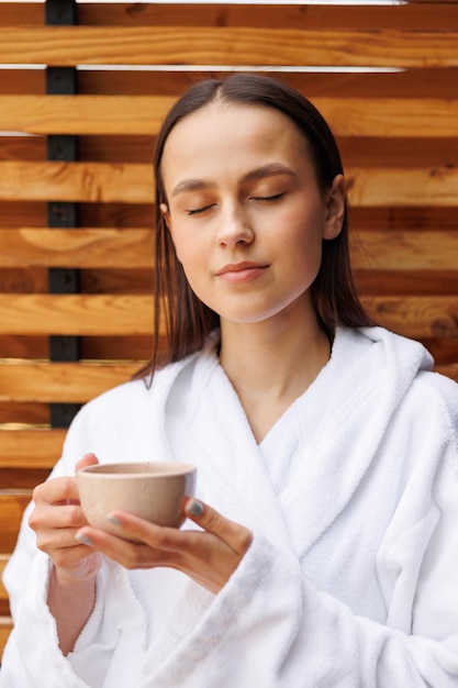 Bastante joven en una bata de baño disfrutando de un delicioso té en la terraza de un hotel spa