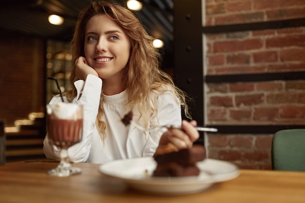 Bastante joven en blusa blanca sentado a la mesa con café caliente y postre dulce