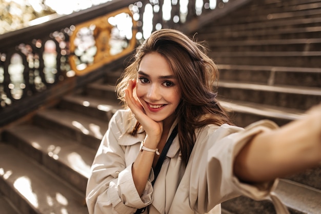 Bastante joven con cabello moreno esponjoso, labios rojos y elegante gabardina sonriendo, posando en la cálida ciudad otoñal y haciendo selfie durante el día