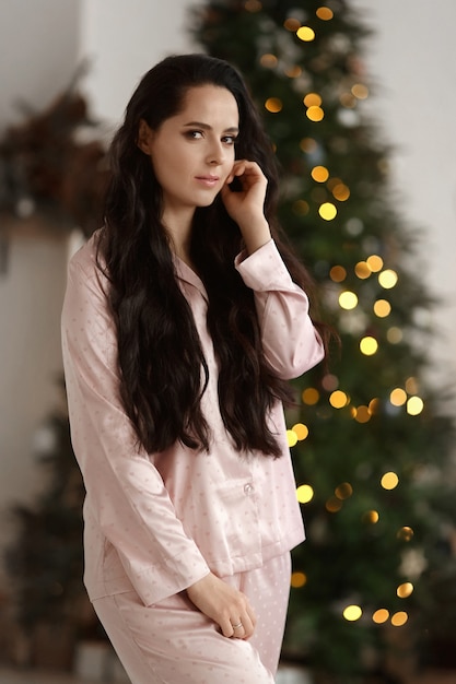 Bastante joven con cabello oscuro vistiendo ropa de dormir de moda posando junto al árbol de Navidad decorado.