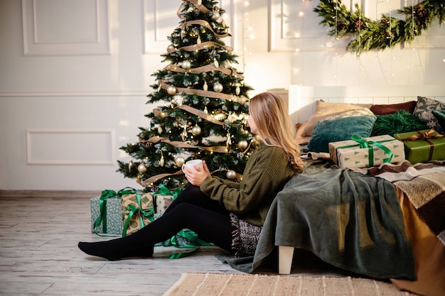 Bastante joven con cabello rubio se sienta cerca de un árbol de Navidad con café. El año nuevo llegará pronto. Ambiente navideño en un hogar acogedor.