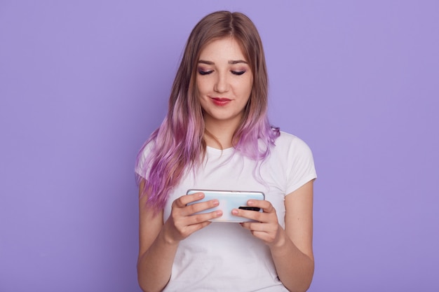 Bastante joven con cabello violeta se viste de camiseta blanca casual con teléfono inteligente en las manos, utilizando Internet inalámbrico para navegar por Internet con aspecto positivo, aislado sobre fondo lila.