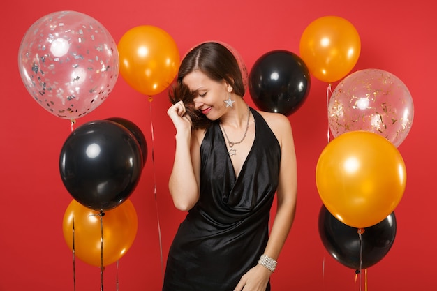 Bastante joven con la cabeza baja en vestido negro celebrando sosteniendo su cabello en globos de aire de fondo rojo brillante. Día internacional de la mujer, feliz año nuevo, concepto de fiesta de vacaciones de maqueta de cumpleaños.