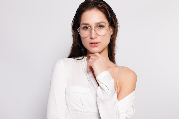 Bastante joven en camiseta blanca en blanco, con gafas, pared vacía, retrato de estudio. Sonriente mujer morena en anteojos