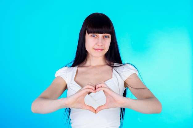 Bastante joven chica de pelo largo haciendo forma de corazón con los dedos, fondo azul claro