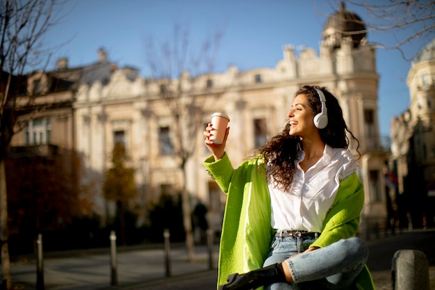 Bastante joven escuchando música con el teléfono inteligente en la calle y sosteniendo café para llevar