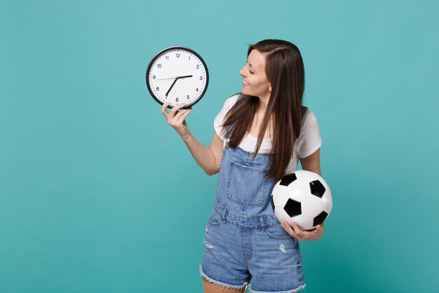 Bastante joven fanático del fútbol anímate a apoyar al equipo favorito con una pelota de fútbol, mirando el reloj redondo aislado en un fondo azul turquesa. Emociones de la gente, concepto de estilo de vida de ocio familiar deportivo.