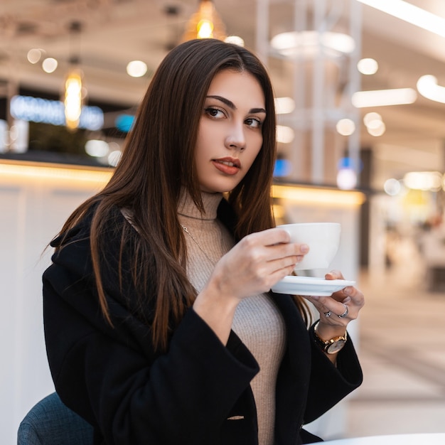 Bastante joven hermosa mujer morena con pelo largo en abrigo negro de moda con taza de café está sentado en la cafetería en el centro comercial. Atractiva chica moderna en ropa de moda disfruta de una bebida caliente en el interior.