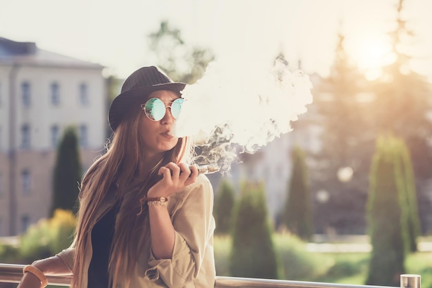 Bastante joven hipster mujer en sombrero negro vape ecig dispositivo de vapeo al atardecer imagen tonificada