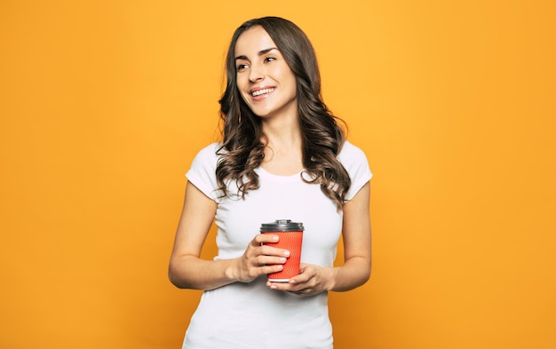 Bastante joven con largo cabello oscuro ondulado y llamativa sonrisa en su rostro sostiene una taza de café sabroso caliente en sus manos