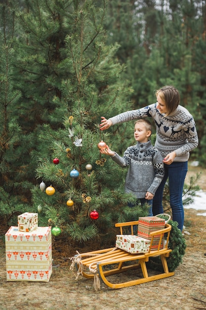 Bastante joven madre y el hijo decorando un árbol de Navidad en el bosque de invierno al aire libre