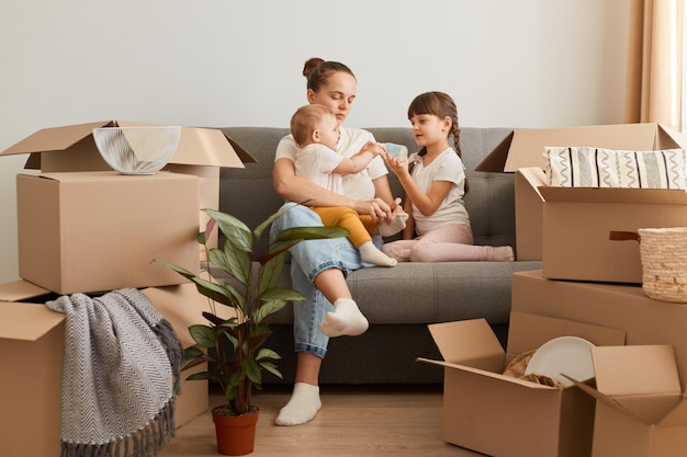 Bastante joven madre y sus lindas hijas pequeñas sentadas en la tos del nuevo apartamento y desempacando cajas de mudanza, expresando emociones positivas, rodeadas de paquetes con pertenencias.