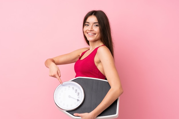 Bastante joven con máquina de pesaje sobre rosa aislado sosteniendo una máquina de pesaje y apuntando