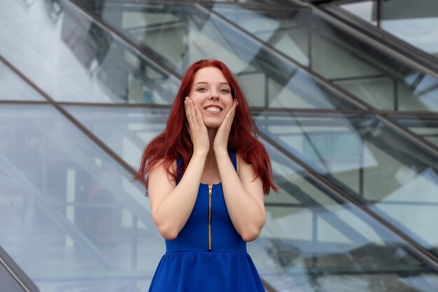 Bastante joven con un maravilloso sentido del humor cabello rojo y un vestido azul en una calle de la ciudad riéndose de la cámara