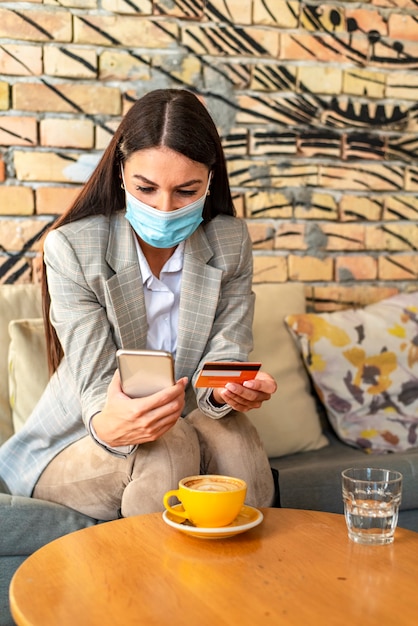 Bastante joven con mascarilla protectora mediante teléfono móvil anc tarjeta de crédito en el café