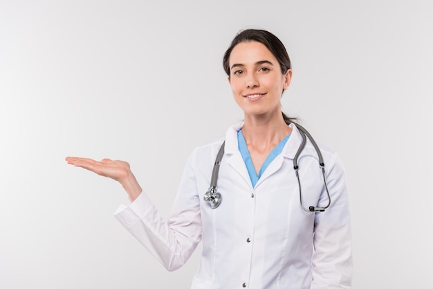 Bastante joven médico sonriente en bata blanca que presenta un nuevo producto médico mientras está de pie frente a la cámara de forma aislada