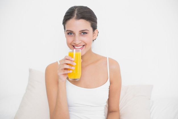 Bastante joven modelo de cabello castaño en pijama blanco bebiendo jugo de naranja