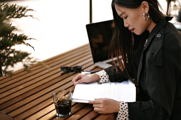 Bastante joven mujer asiática bronceada en elegante gabardina negra posa afuera con un vaso de café y lee un libro