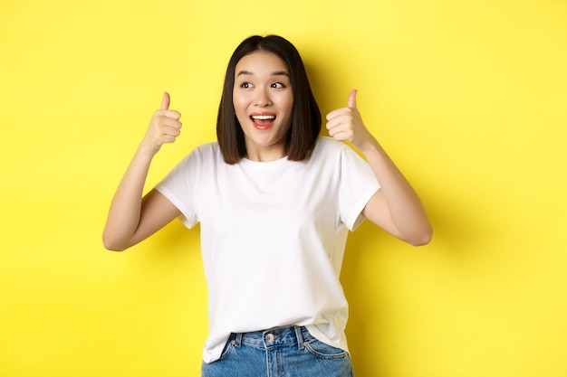 Bastante joven mujer asiática en camiseta blanca, mostrando los pulgares hacia arriba y sonriendo, elogia la buena oferta, recomienda el producto, de pie satisfecha sobre fondo amarillo.