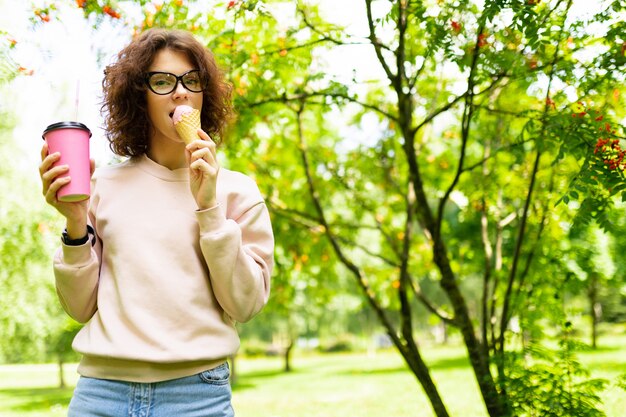 Bastante joven mujer caucásica ir a dar un paseo en el parque, bebe café y come helado