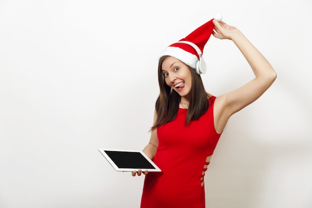 Bastante joven mujer feliz en vestido rojo y sombrero de Navidad escuchando música en auriculares, sosteniendo la tableta sobre fondo blanco. Gadget de niña Santa aislado. Vacaciones de año nuevo 2018. Anuncio de espacio de copia.