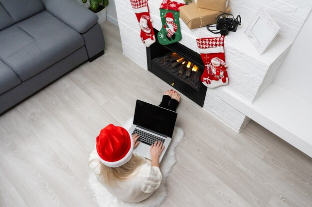 Bastante joven mujer de moda de invierno sentado en el suelo sosteniendo el gadget portátil en frente de la decoración de Navidad, mientras mira a la cámara.