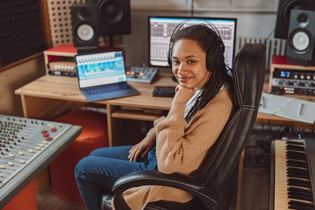 Bastante joven mujer multiétnica músico compositor ingeniero de sonido en el trabajo en el estudio de grabación sonríe mirando a la cámara