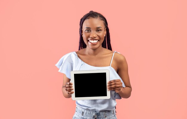 Bastante joven mujer negra sosteniendo tableta con pantalla vacía en maqueta de fondo de estudio rosa