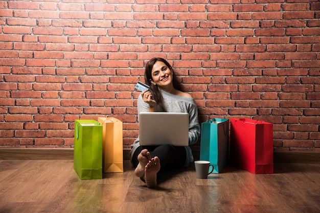 Bastante joven mujer o niña india de compras con tarjeta de crédito o débito y un portátil mientras está sentado sobre un piso de madera contra la pared de ladrillo rojo