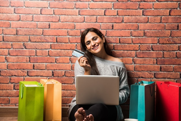 Bastante joven mujer o niña india de compras con tarjeta de crédito o débito y un portátil mientras está sentado sobre un piso de madera contra la pared de ladrillo rojo