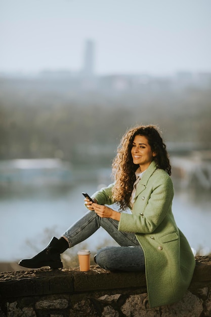 Bastante joven mujer de pelo rizado con smartphone mientras está sentado junto al río y bebiendo café para llevar