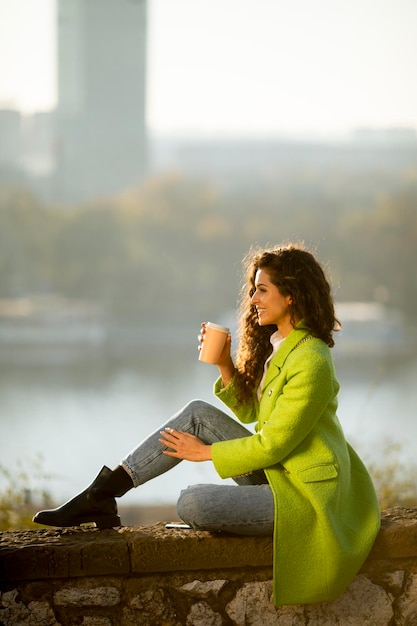 Bastante joven mujer de pelo rizado con smartphone mientras está sentado junto al río y bebiendo café para llevar