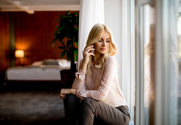 Bastante joven mujer rubia con teléfono móvil junto a la ventana en la habitación