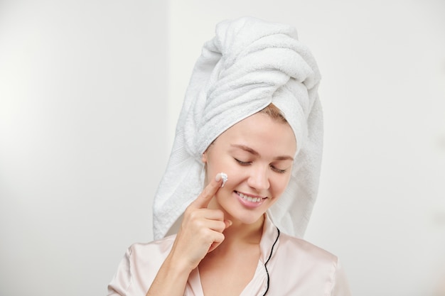 Foto bastante joven mujer satisfecha aplicando crema hidratante en la cara después de los procedimientos de higiene matutinos