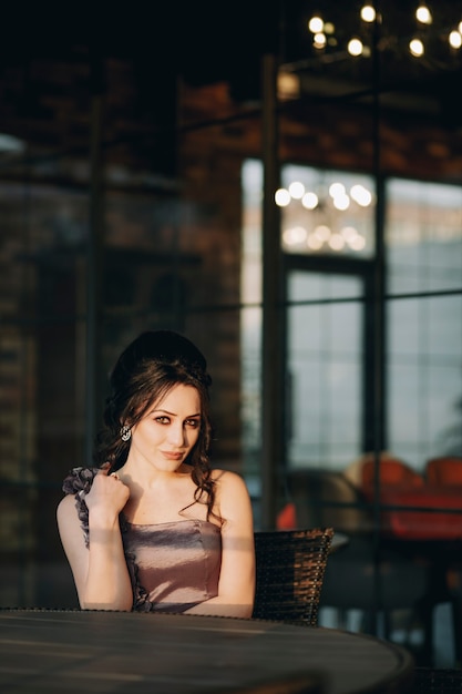 Bastante joven mujer sentada en un café al aire libre frente a ventanas de vidrio y posando para la cámara